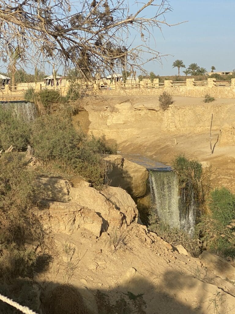 Ein kleiner, natürlicher Wasserfall fließt im Hochformat über sandfarbene Felsen und Klippen in der Oase Fayyum, umgeben von kargem Gebüsch und Palmen im Hintergrund.