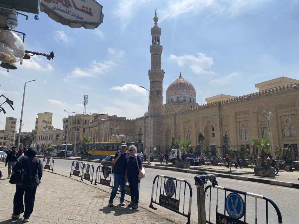 Anne und Ulrich stehen auf einem Gehweg in Kairo vor der Sayyeda-Zeinab-Moschee mit ihrem hohen Minarett und der verzierten Kuppel unter leicht bewölktem Himmel.