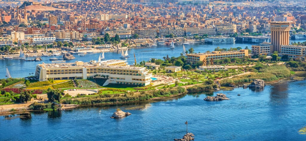 Eine Panorama-Luftaufnahme einer grünen Nilinsel mit Hotelanlagen in Aswan, dahinter der Fluss mit Segelbooten und die dicht bebaute Stadt im Hintergrund.