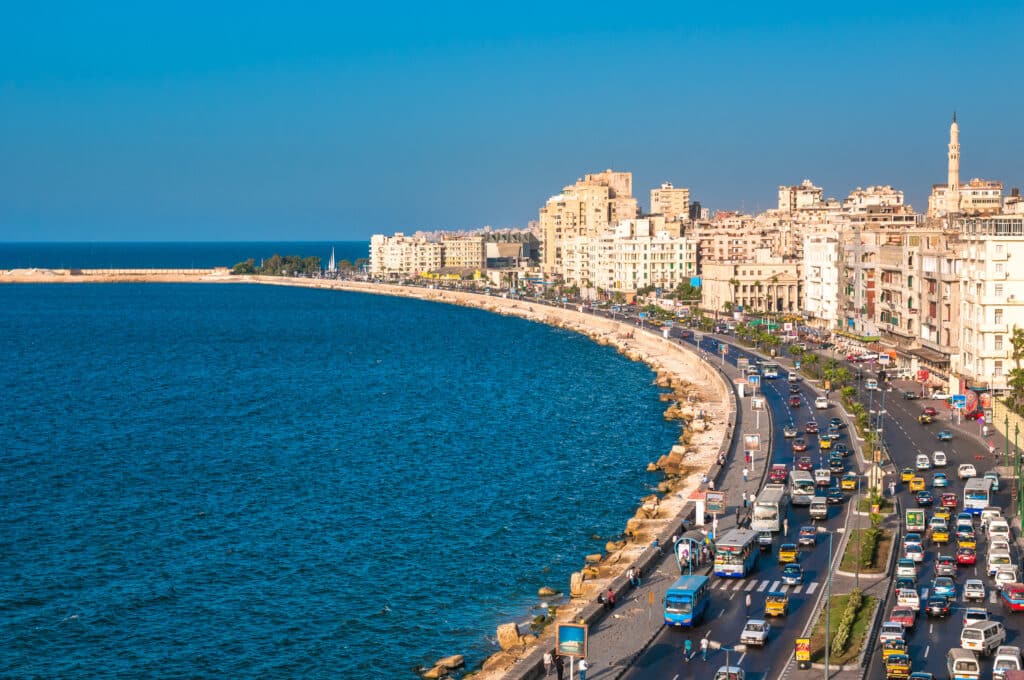 Blick von oben auf die geschwungene Corniche-Uferpromenade von Alexandria mit dichtem Autoverkehr direkt neben dem blauen Mittelmeer.
