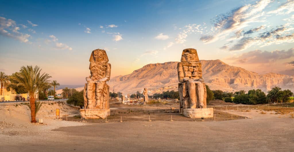 Die beiden riesigen Memnon-Kolosse stehen imposant in einer Wüstenlandschaft vor den Bergen von Theben-West in Luxor.