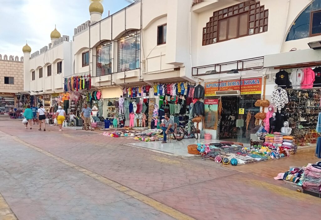 Eine breite Fußgängerzone im Old Market von Sharm el Sheikh mit zweistöckigen, hellen Gebäuden, vor denen Kleidung, bunte Souvenirs und Textilien ausgestellt sind.