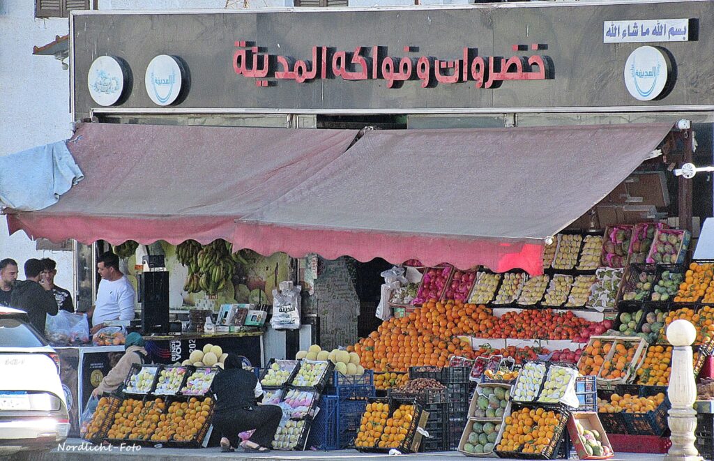 Ein farbenfroher, gut sortierter Obst- und Gemüseladen in Hay el Nour, Sharm el Sheikh, mit großen Kisten voller Orangen, Bananen und Mangos unter einer roten Markise.